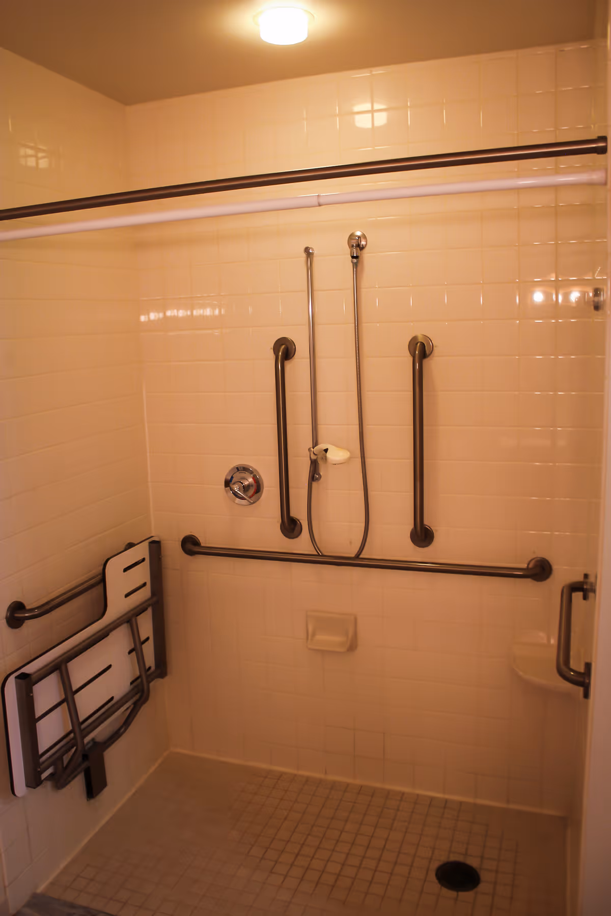 A tiled shower area with multiple grab bars for accessibility, a handheld showerhead, a foldable shower seat attached to the wall, and a soap holder. The shower floor has small square tiles and a drain, and there is a ceiling light above.