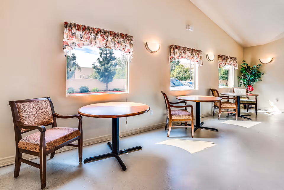 Bright communal dining area with round tables and upholstered chairs positioned by three windows.