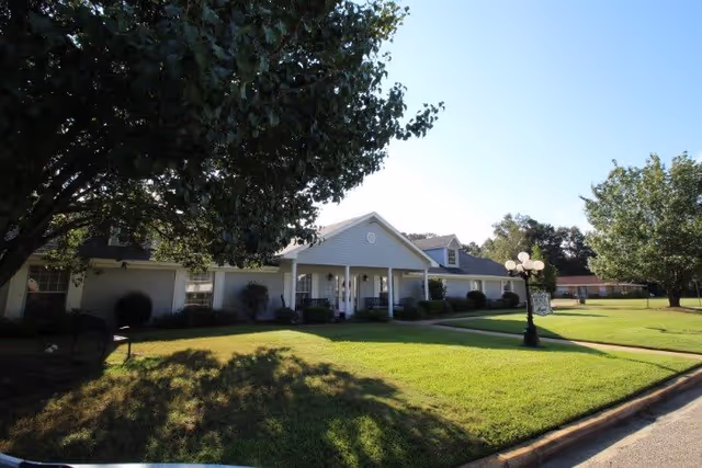 Exterior view of a single-story senior living facility building with a white facade, a covered entrance with columns, and a well-maintained lawn with trees and a street lamp in front.
