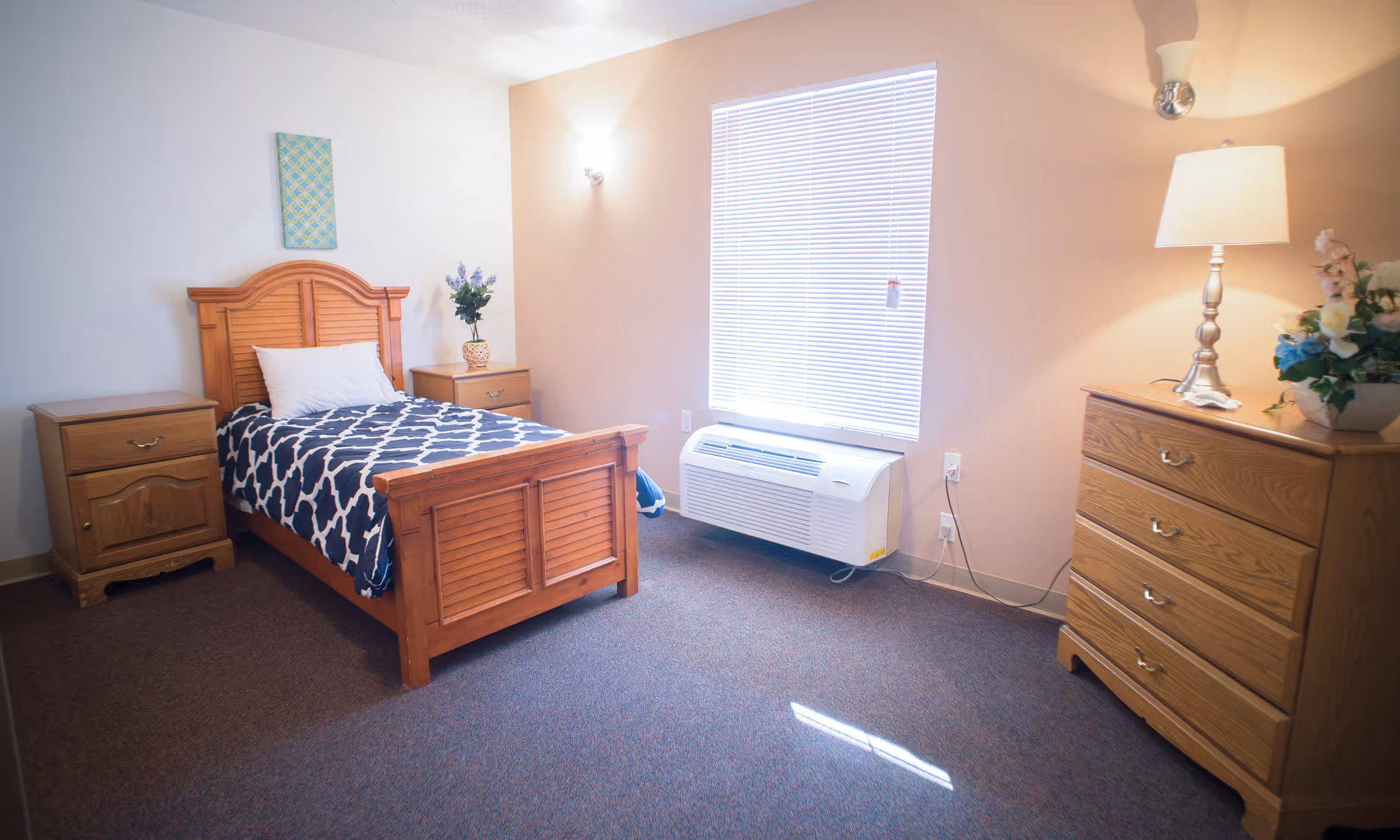 Sunlit furnished bedroom with a wooden twin bed, matching nightstands and dresser, a lamp, and a window with blinds.