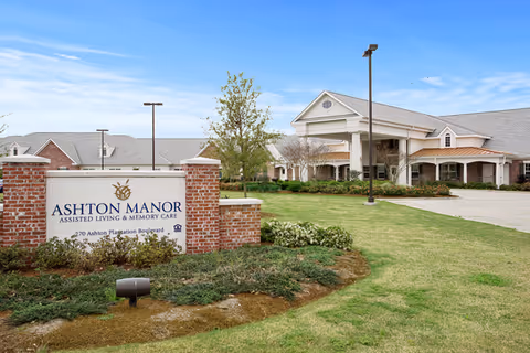 Exterior view of Ashton Manor assisted living and memory care facility showing the main building with a covered entrance, surrounded by landscaped greenery and a clear blue sky. A large sign in front reads 'Ashton Manor Assisted Living & Memory Care, 270 Ashton Plantation Boulevard.'