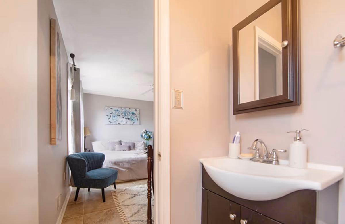 View of a bedroom with a bed, blue armchair, and floral wall art, seen through an open doorway from a small bathroom with a sink, mirror, and soap dispenser.