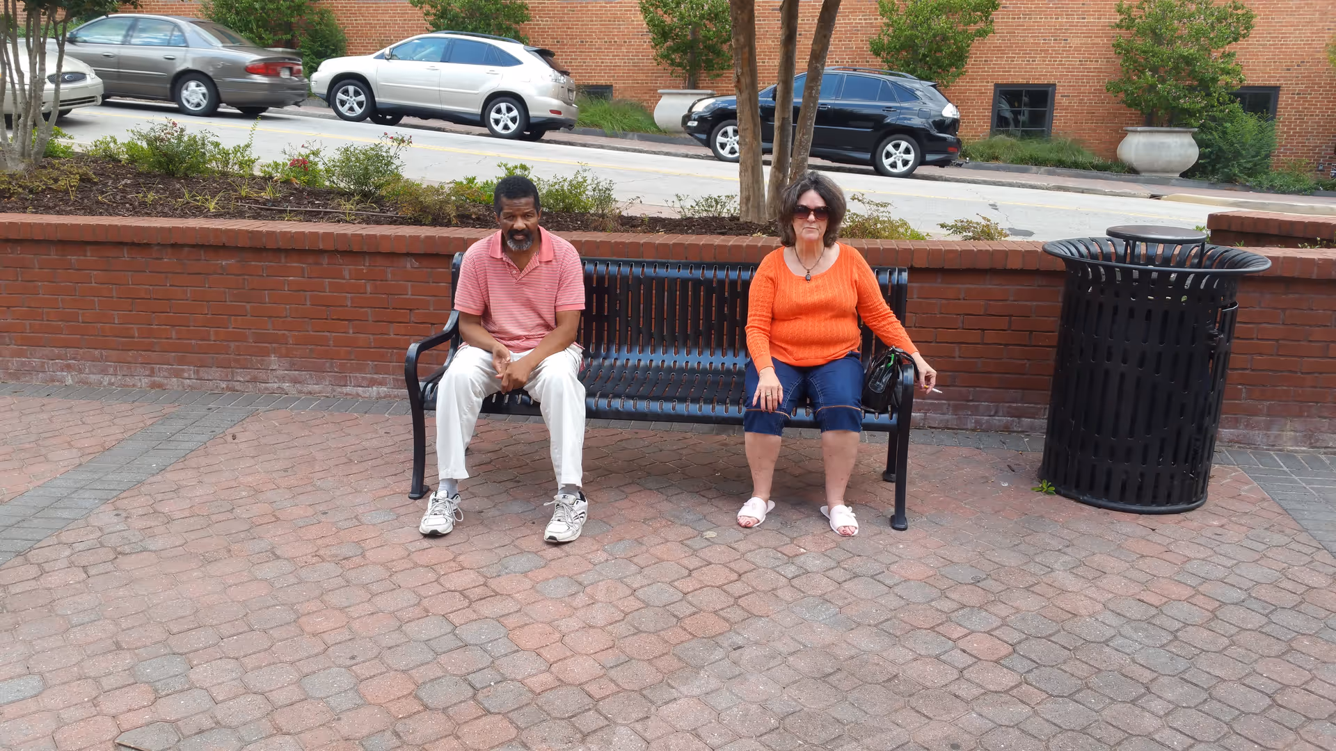 Two people sitting on a black metal bench on a brick-paved sidewalk. Behind them is a low red brick wall with some plants and trees, and a street with parked cars. A large black trash can is next to the bench on the right.