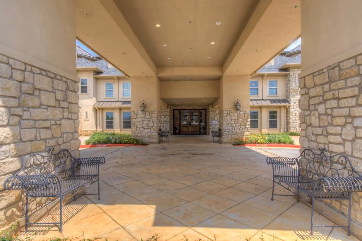 Covered entrance area of a senior living facility with stone pillars and beige walls, featuring two black metal benches on either side and a double glass door entrance in the background.