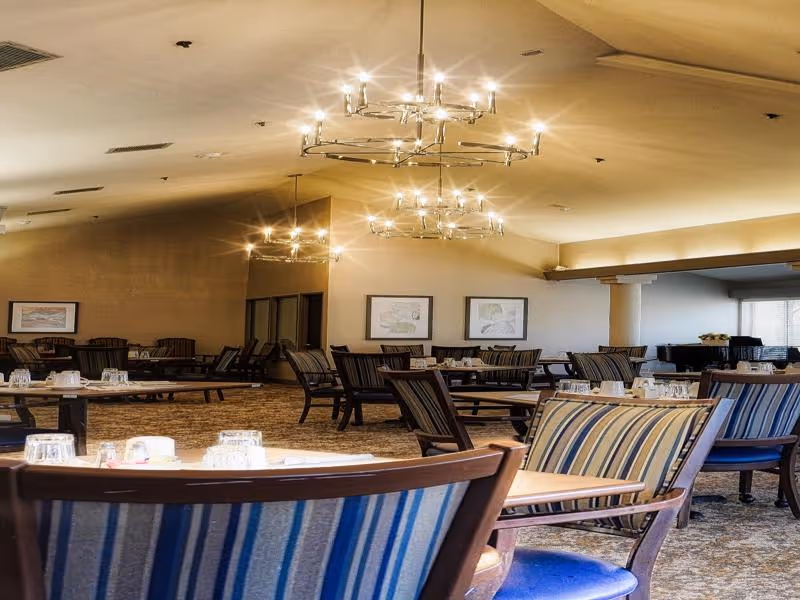 A spacious dining room with multiple wooden tables and chairs, each table set with glasses and napkins. The room features striped upholstered chairs, warm lighting from modern chandeliers, framed artwork on the walls, and large windows allowing natural light.