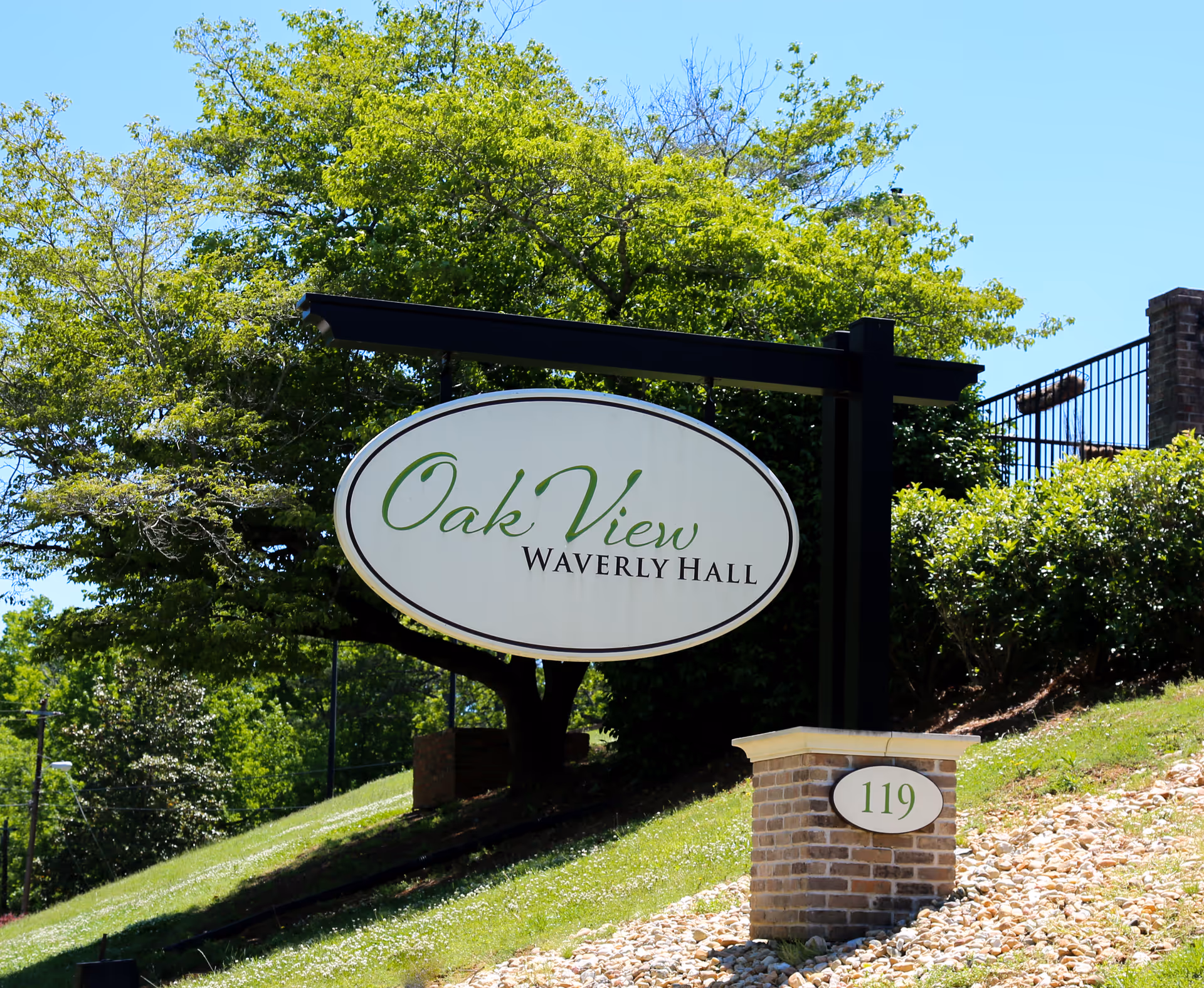 Outdoor view of a sign for Oak View Waverly Hall mounted on a black metal frame with greenery and trees in the background. A brick pillar with the number 119 is visible next to the sign.