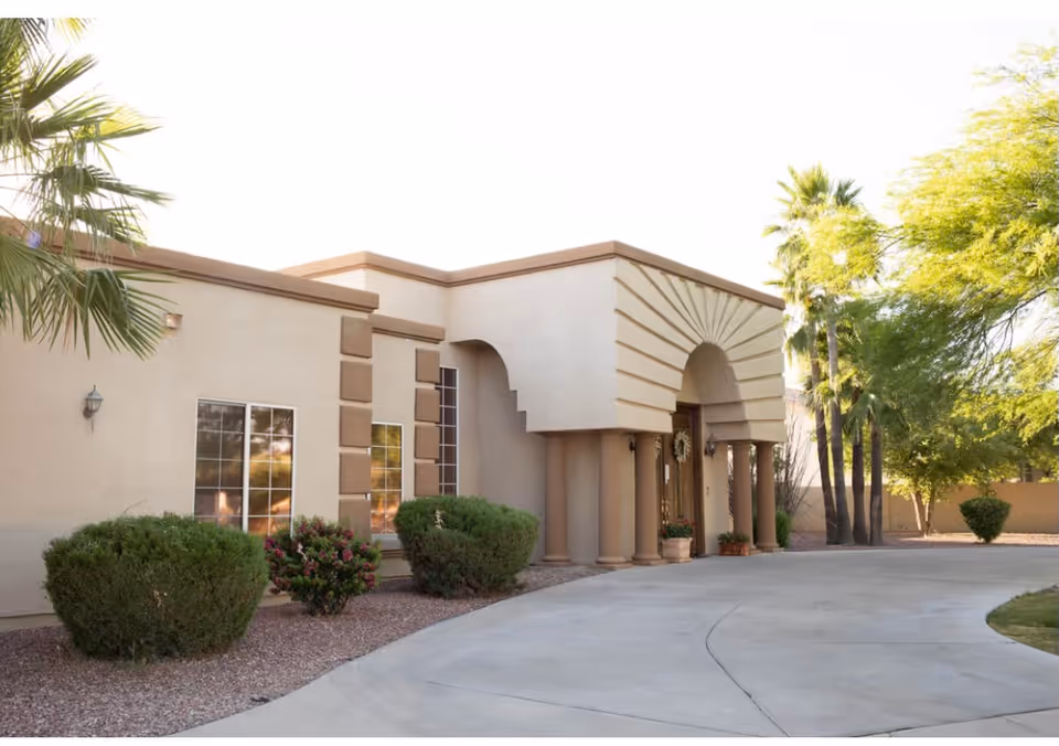 Exterior view of Golden Swan Manor Assisted Living facility showing a beige building with decorative columns and an arched entrance, surrounded by bushes, palm trees, and a curved driveway.