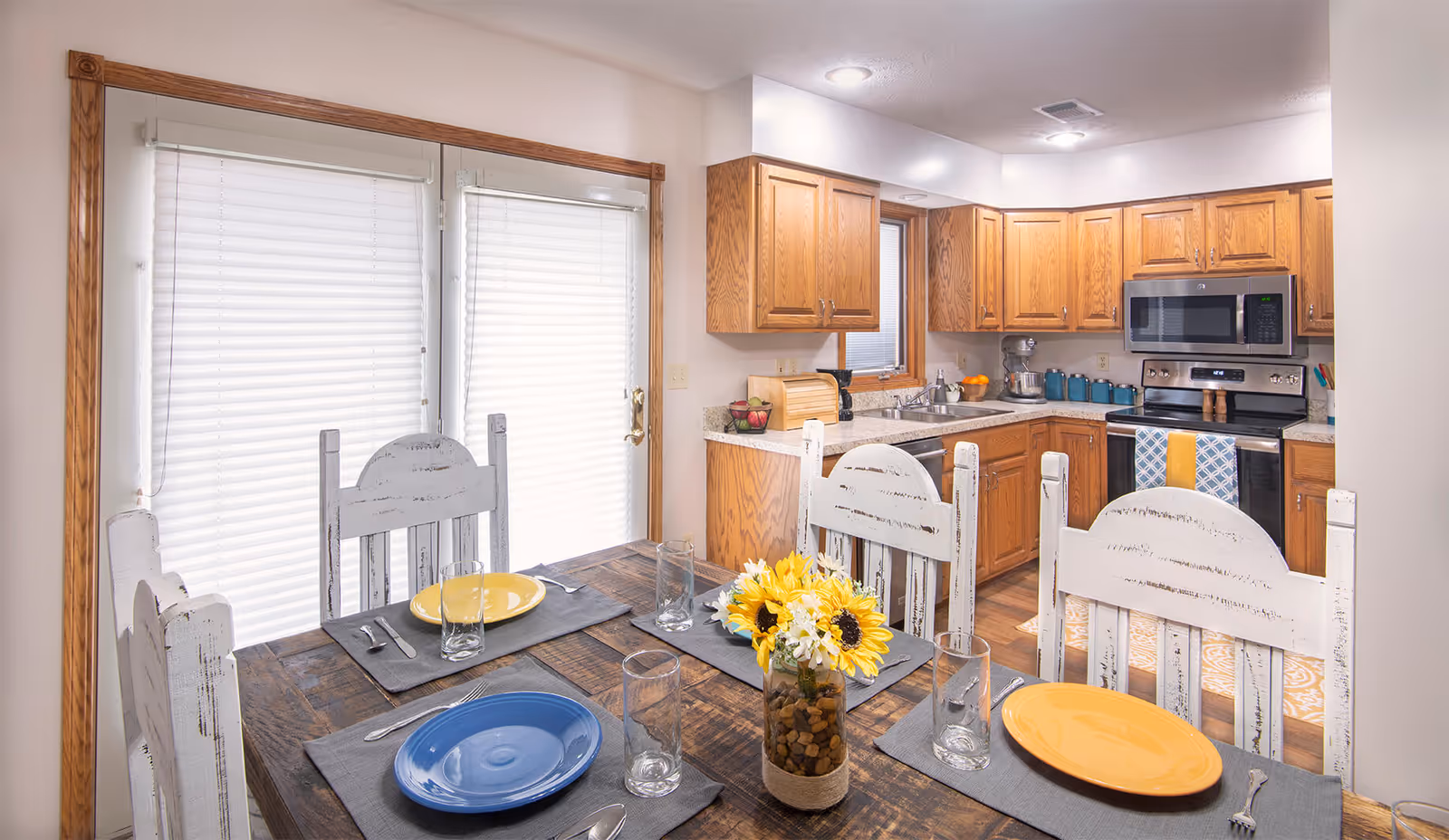 Sunlit dining area with a rustic wooden table set with colorful plates and a vase of sunflowers, opening into a kitchen with oak cabinets and stainless steel appliances.