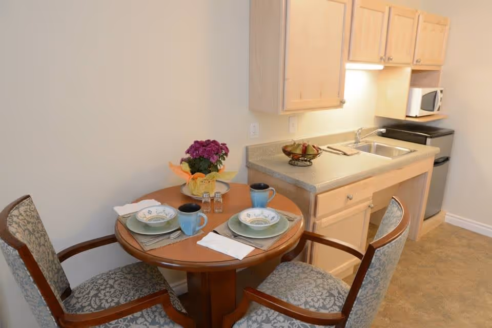 A small dining area with a round wooden table set for two, featuring plates, bowls, mugs, napkins, and salt and pepper shakers. A potted purple flower centerpiece is on the table. Adjacent to the table is a compact kitchen area with light wood cabinets, a countertop with a fruit bowl, a sink, a microwave, and a small refrigerator.
