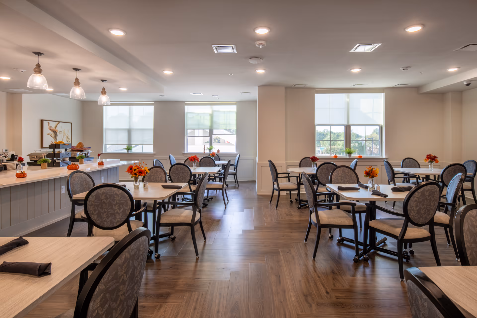 A bright and spacious dining room with multiple tables and cushioned chairs arranged neatly. Each table has a small floral centerpiece and folded black napkins. Large windows allow natural light to fill the room, and pendant lights hang from the ceiling above a counter area on the left side. The floor is wooden, and the overall atmosphere is clean and inviting.