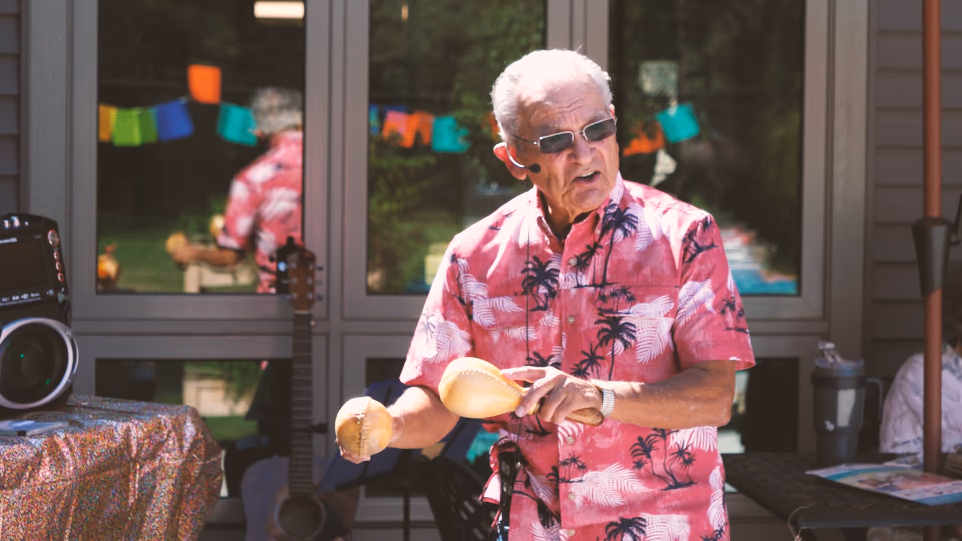 An elderly man wearing sunglasses and a pink Hawaiian shirt with palm tree patterns is playing maracas outdoors. Behind him, there is a window reflecting colorful decorations and a guitar leaning against the wall. A speaker and a table with a patterned cloth are also visible nearby.