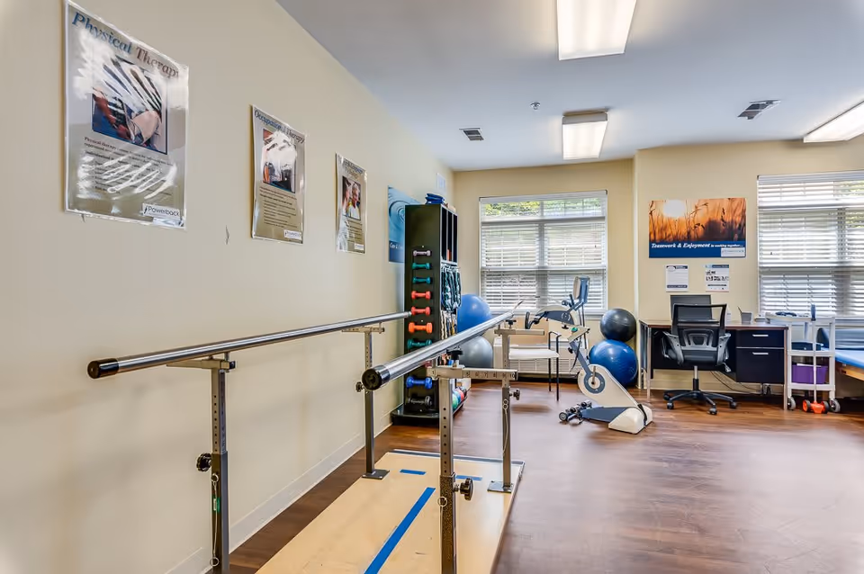 A physical therapy room with parallel bars for walking exercises, exercise balls, a stationary bike, dumbbells, and a desk with a chair. Posters related to physical and occupational therapy are displayed on the walls. The room has large windows with blinds and wood flooring.