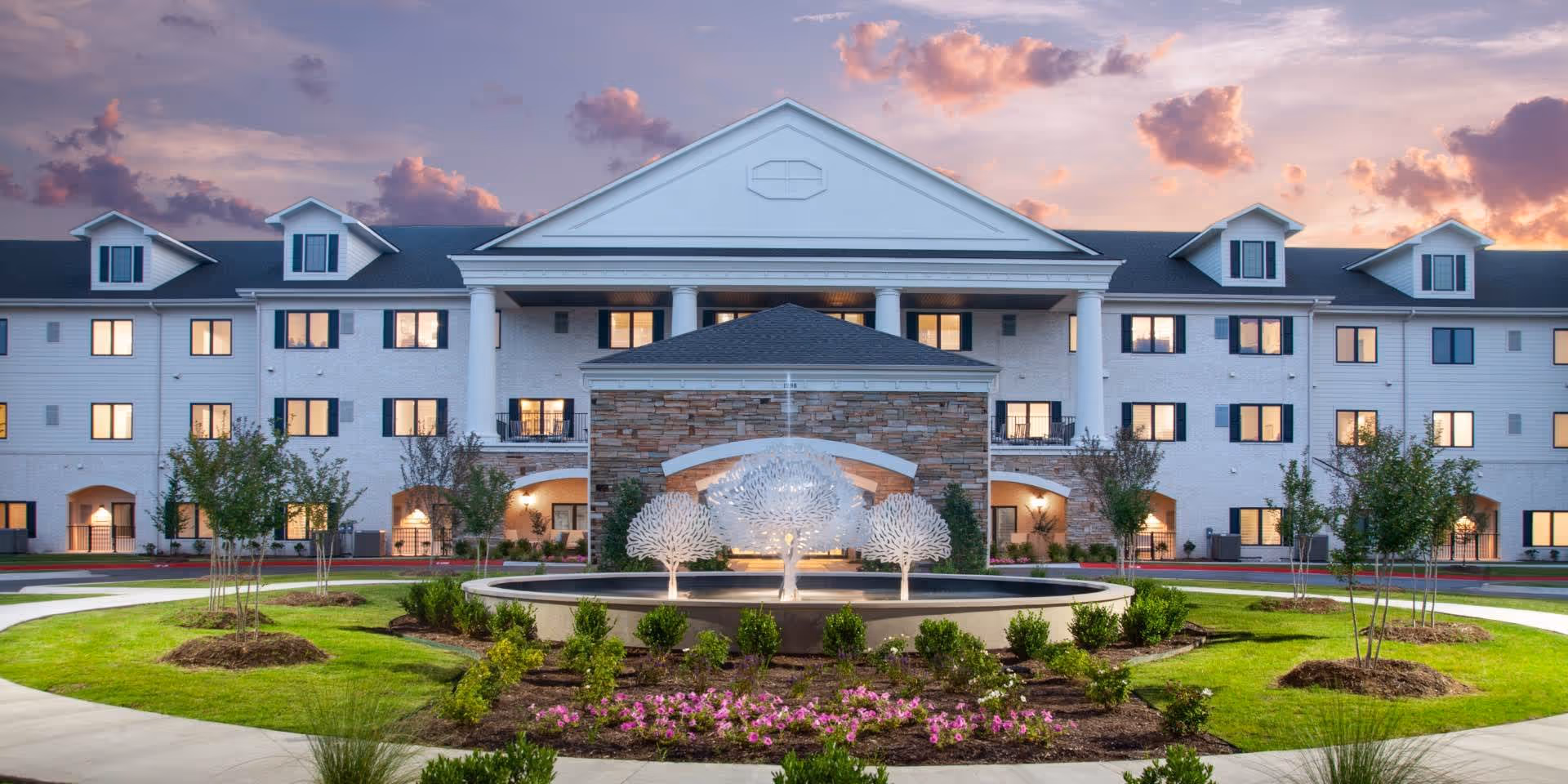 Front exterior of the Grand Village at Clear Creek building with a landscaped circular driveway and illuminated decorative fountain at sunset.