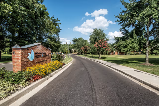 A paved road leading into a senior living facility with a brick sign on the left that reads Memphis Jewish Home & Rehab, surrounded by well-maintained landscaping including yellow flowers and green shrubs. Trees and a grassy lawn line both sides of the road under a partly cloudy blue sky.