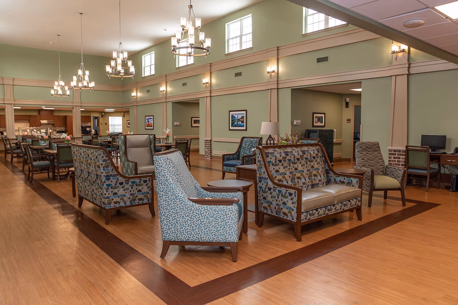 A spacious, well-lit common area in a senior living facility featuring multiple seating arrangements with patterned armchairs and sofas, wooden flooring with a darker wood border, green walls with beige trim, several chandeliers hanging from the ceiling, and a dining area with tables and chairs in the background.