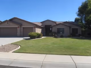 Single-story residential building with a three-car garage, beige exterior walls, and a well-maintained green lawn in front under a clear blue sky.