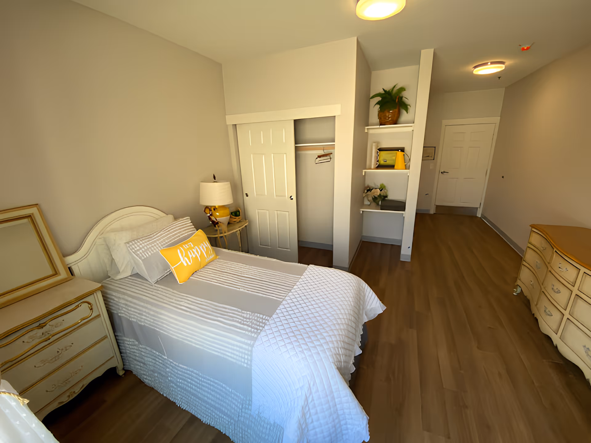 A cozy bedroom with a single bed covered in gray and white striped bedding and a yellow pillow that says 'happy'. The room features a wooden dresser with a mirror, a small side table with a yellow lamp, an open closet with hangers, built-in shelves with decorative items, and a wooden floor. The walls are painted light beige, and there is a white door at the end of the room.