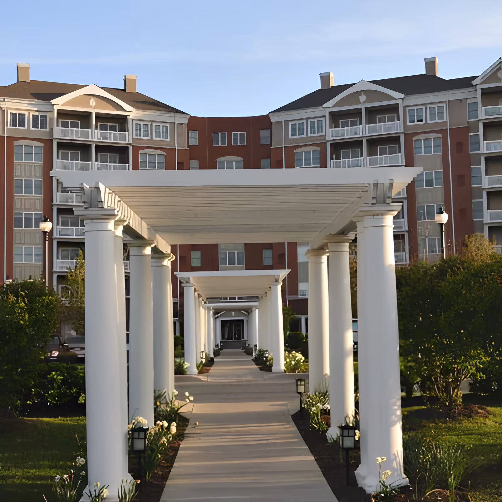 A long covered pergola walkway with white columns leading to the entrance of a multi-story residential building.