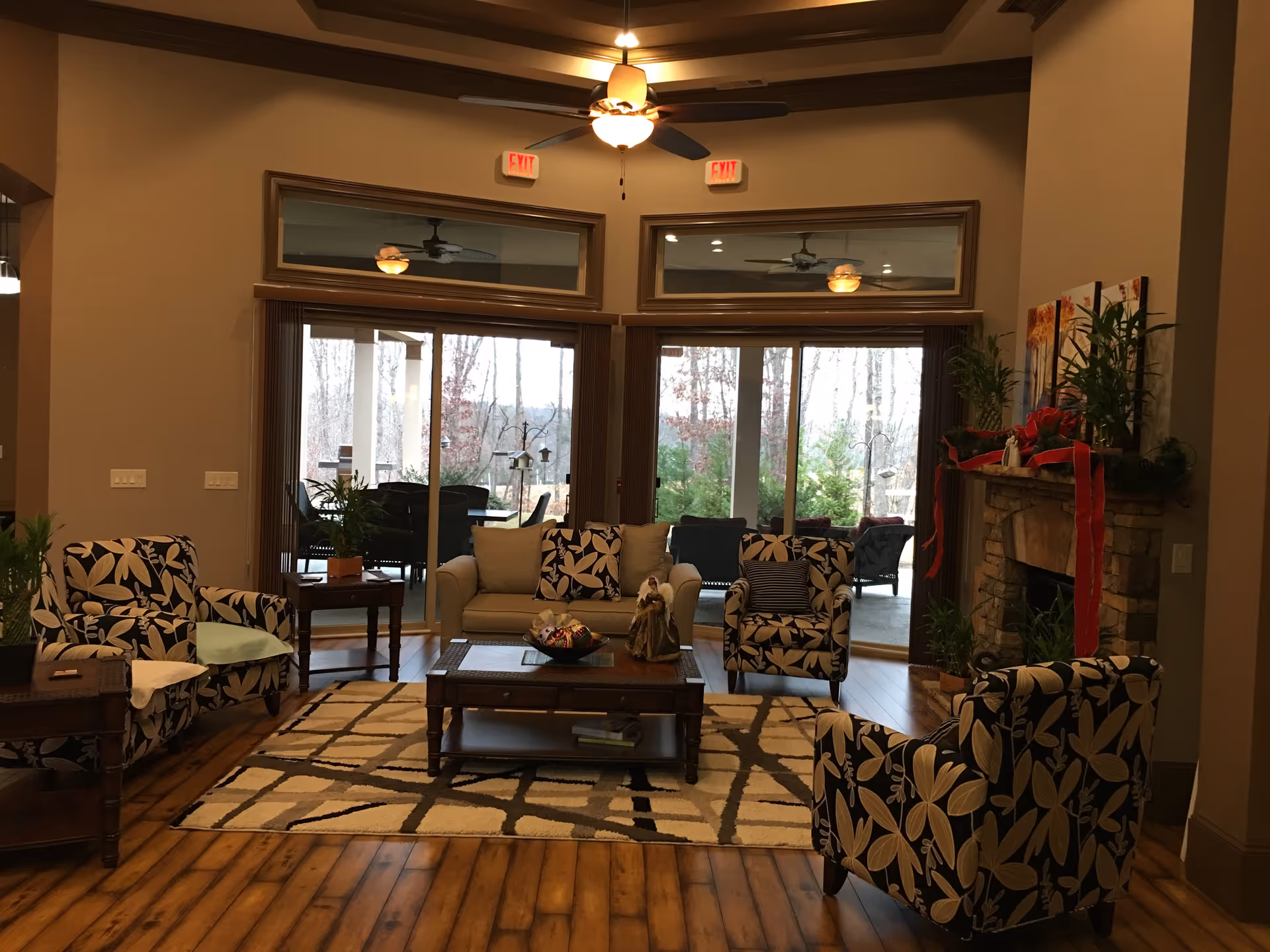 A cozy senior living community lounge with floral patterned armchairs, a beige sofa, a wooden coffee table on a patterned rug, and a stone fireplace decorated with plants and red ribbons. Large windows and glass doors provide a view of the outdoor patio and trees beyond.