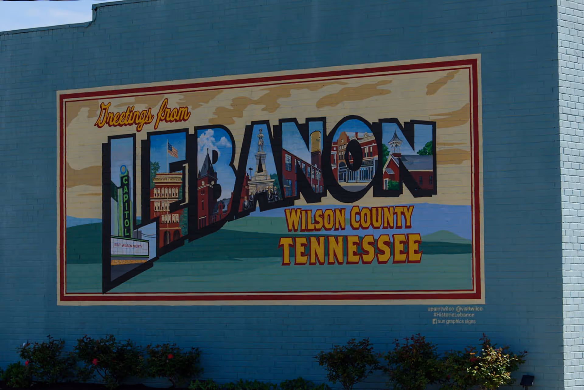 A mural painted on a blue brick wall that says 'Greetings from LEBANON WILSON COUNTY TENNESSEE' with each letter of LEBANON containing images of local landmarks and buildings. There are small bushes and plants at the base of the wall.