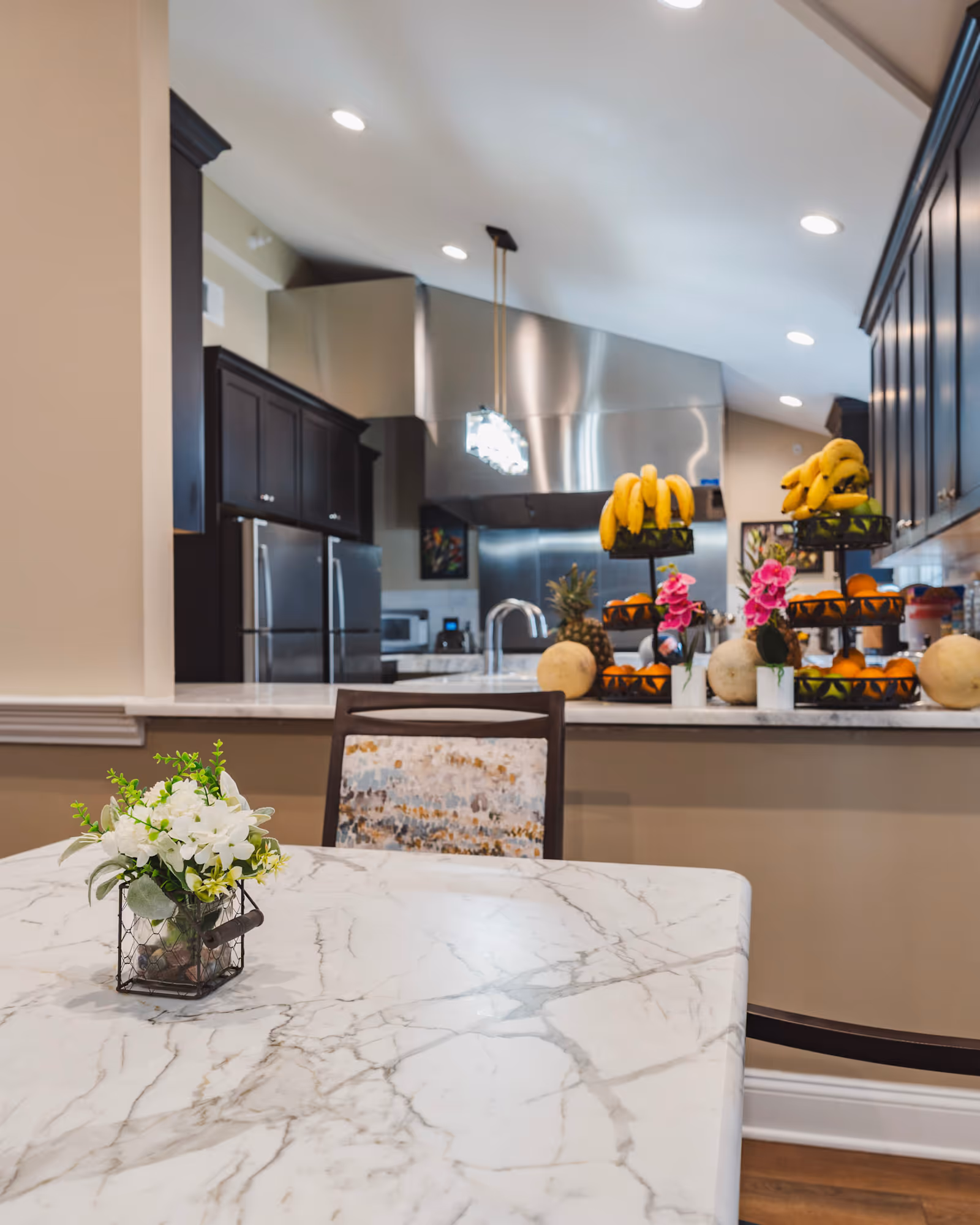Marble dining table with a small floral centerpiece in front of an open kitchen with stainless-steel appliances and tiered fruit displays.