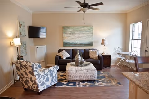 Living room with a dark sofa, patterned armchair and ottoman, wall art, ceiling fan, and a small wall-mounted TV.