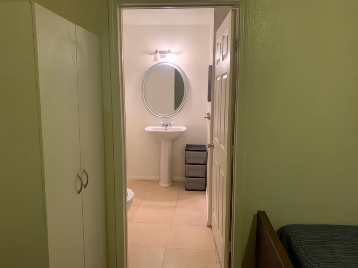 View through an open door into a bathroom with a white pedestal sink, an oval mirror above it, a light fixture on the wall, and a small black storage drawer unit next to the sink. Part of a bed and a white closet with double doors are visible in the foreground.