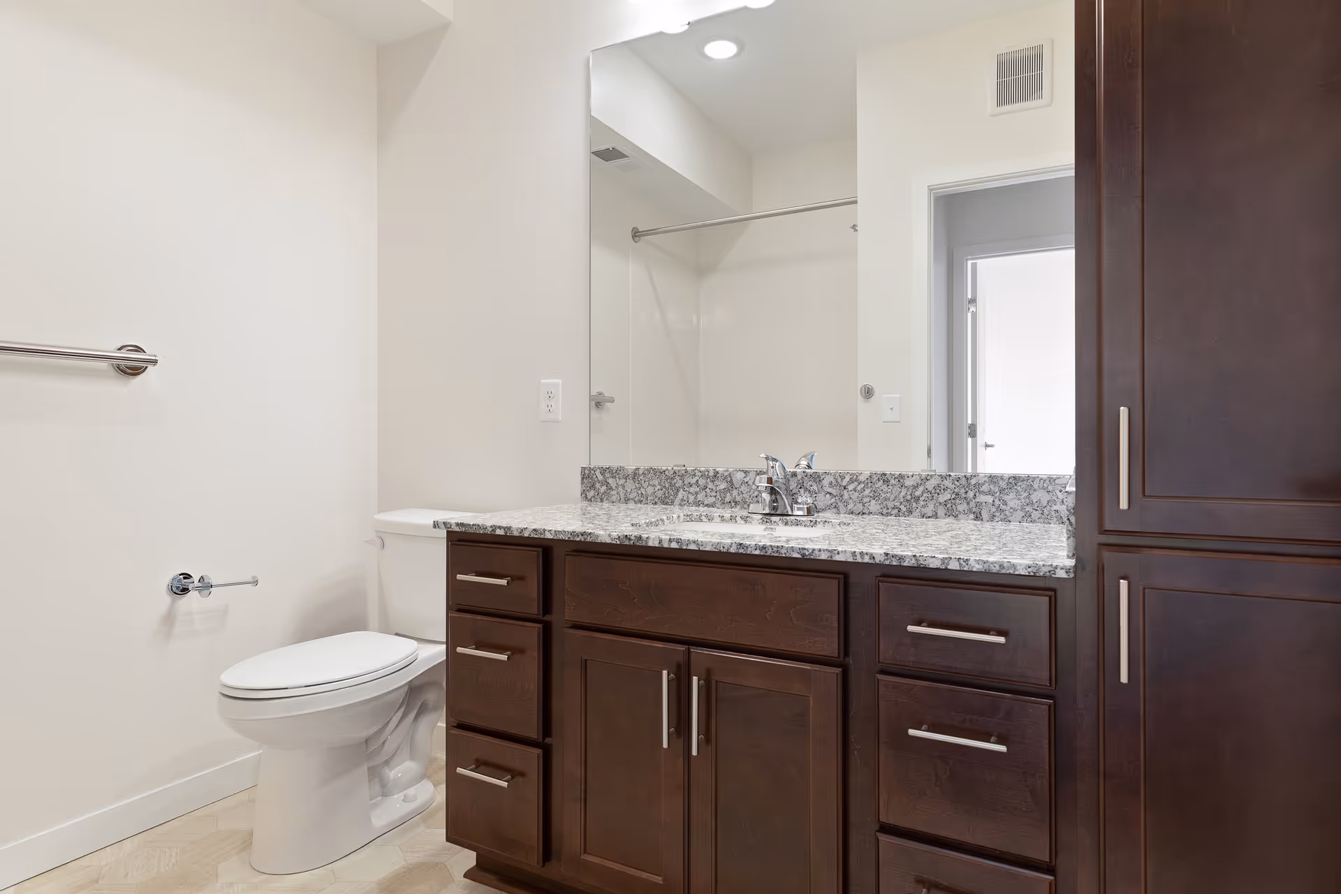 A clean bathroom featuring a white toilet, a granite countertop with a sink, dark wooden cabinets and drawers, a large mirror above the sink, and a towel rack on the wall. The walls are painted white and there is a shower area visible in the mirror's reflection.