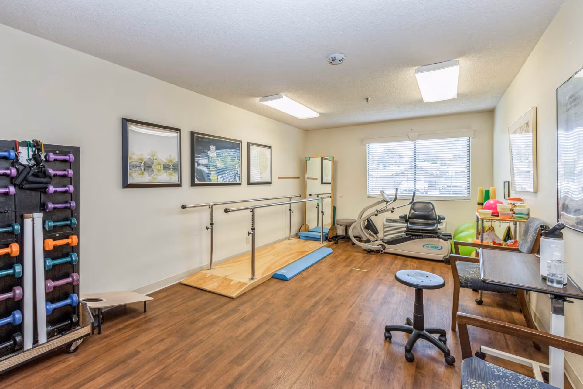 A small exercise room with wooden flooring, featuring a set of parallel bars for walking exercises, a stationary recumbent bike, a rack with colorful dumbbells, exercise balls, and a mirror on the wall. There are framed pictures on the walls and a window with blinds letting in natural light.