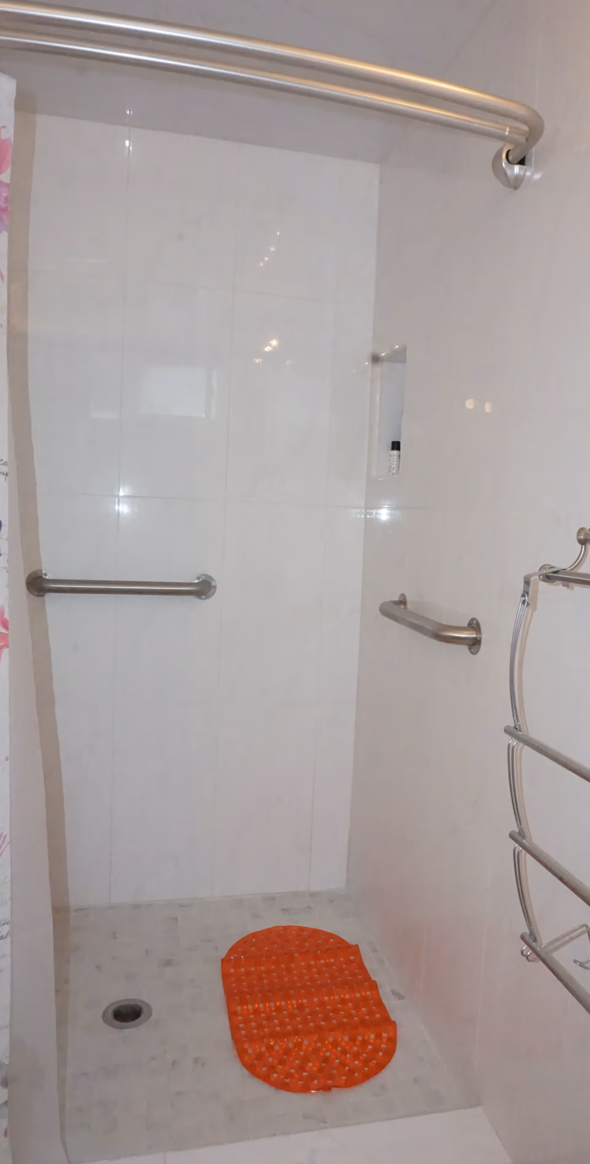 A walk-in shower with white tiled walls and floor, equipped with stainless steel grab bars on the side walls. There is an orange non-slip mat on the shower floor and a curved shower curtain rod at the top.