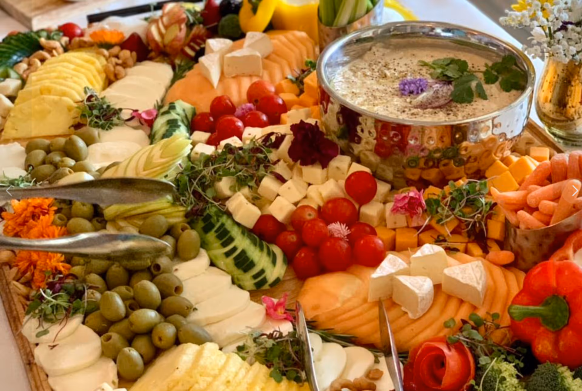 A close-up view of a colorful charcuterie board featuring a variety of sliced fruits, cheeses, cherry tomatoes, olives, baby carrots, and a bowl of creamy dip garnished with herbs and edible flowers.
