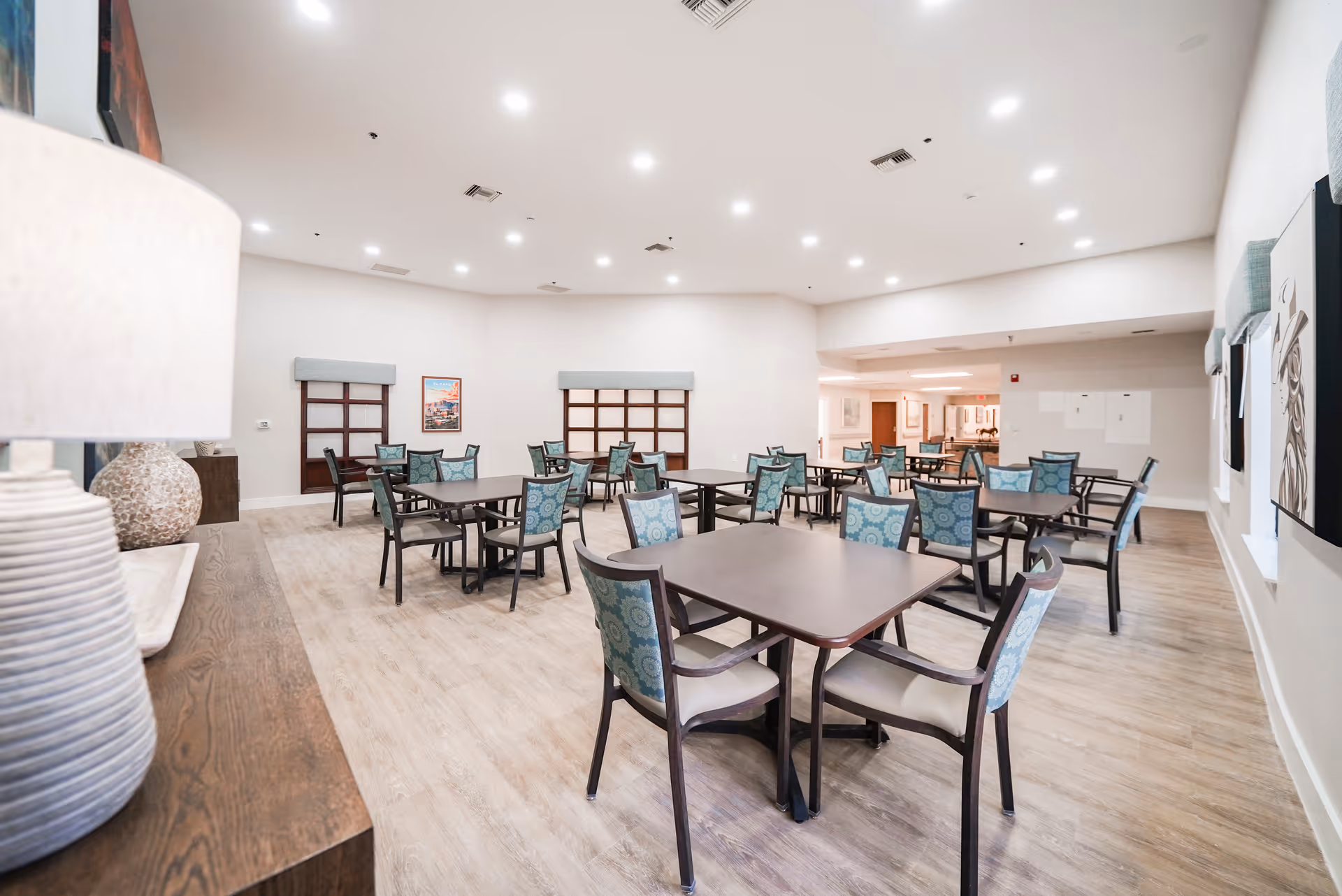 A spacious dining room with multiple square tables and chairs arranged neatly. The chairs have patterned blue upholstery. The room has light-colored walls and wood flooring, with recessed ceiling lights providing bright illumination. Decorative items including a lamp and vases are visible on a wooden sideboard to the left.