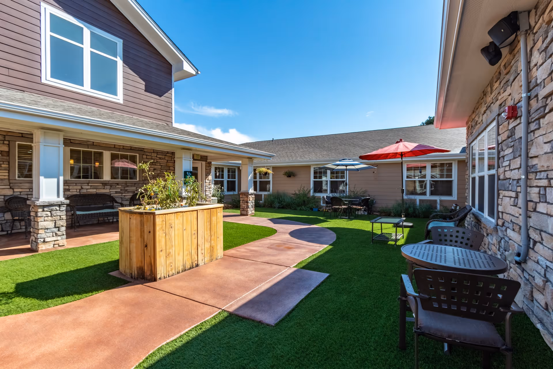 Outdoor courtyard area of a senior living facility with artificial grass, a wooden planter box, several tables with umbrellas, chairs, and stone and siding exterior walls under a clear blue sky.