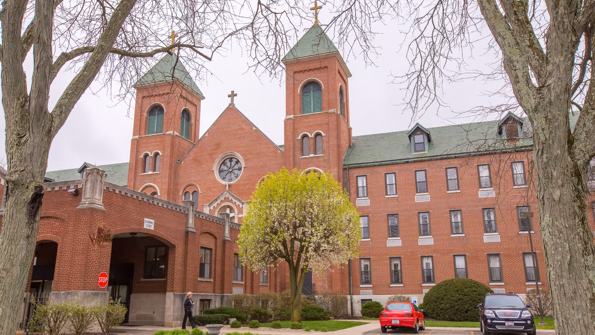 Exterior view of a large red brick building with two tall towers topped with crosses, a central rose window, and a green roof. There is a tree with light green leaves in front of the building, along with some parked cars and a person walking near the entrance. The sky is overcast.