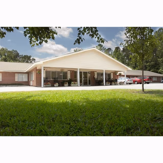 Front exterior view of a single-story brick building with a covered entrance supported by white columns. There are potted plants and rocking chairs on the porch, a green lawn in the foreground, and several cars parked to the right. Trees and a partly cloudy sky are visible in the background.