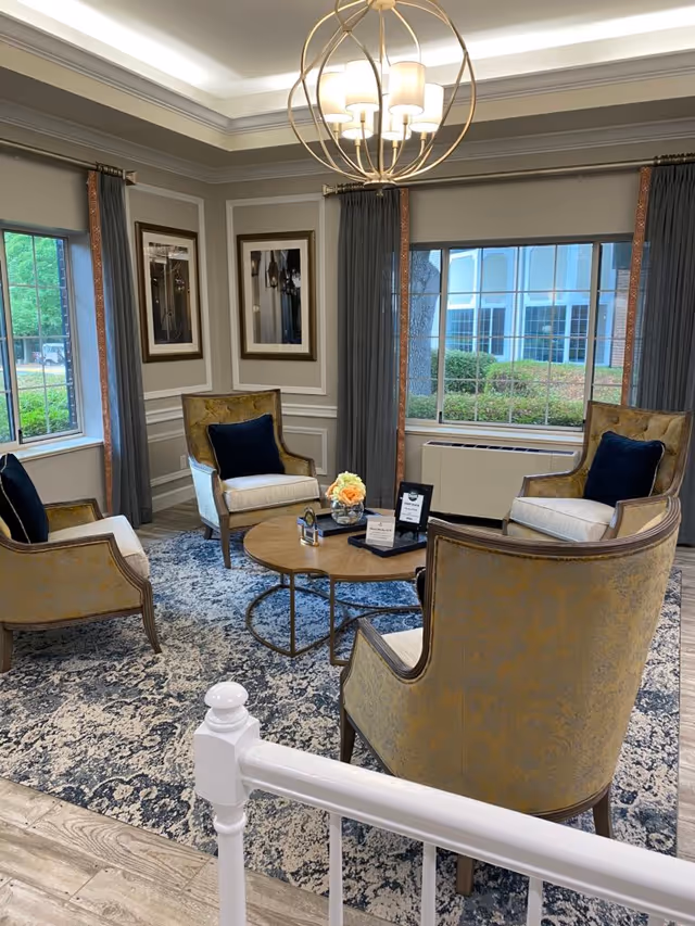 A cozy sitting area with four upholstered armchairs arranged around a round wooden coffee table on a patterned rug. The room has large windows with gray curtains, framed artwork on the walls, and a modern chandelier hanging from the ceiling. Outside the windows, greenery and a building are visible.