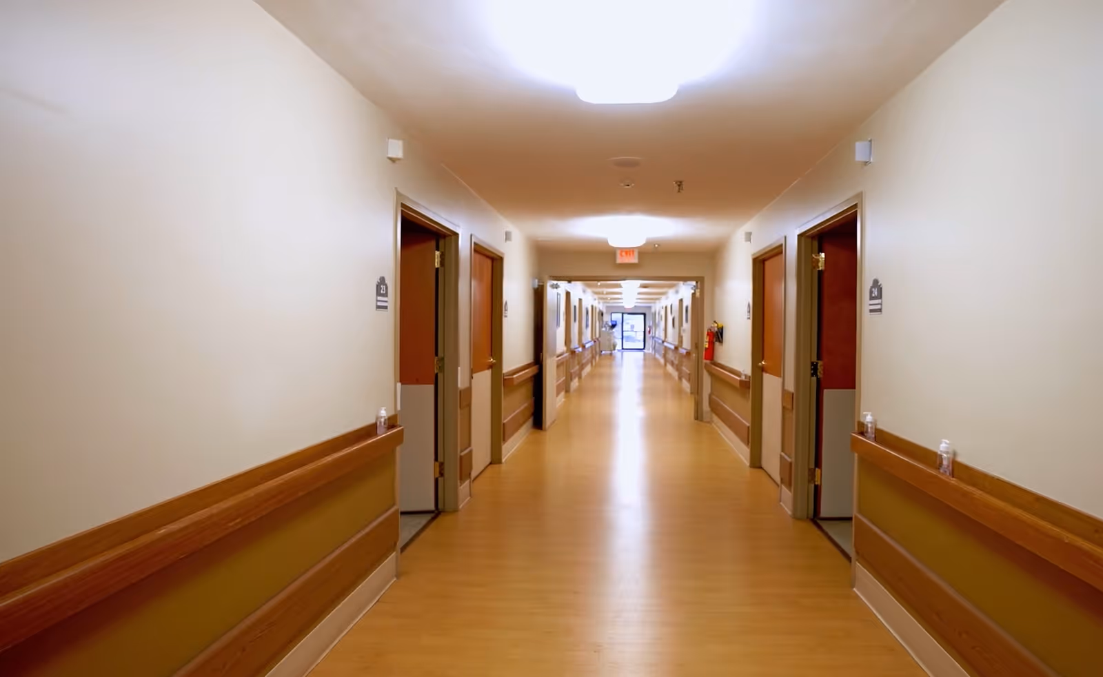 A long, well-lit hallway in a healthcare facility with wooden handrails on both sides and multiple open doors leading to rooms. The floor is light wood, and there is an exit sign visible at the far end of the corridor.