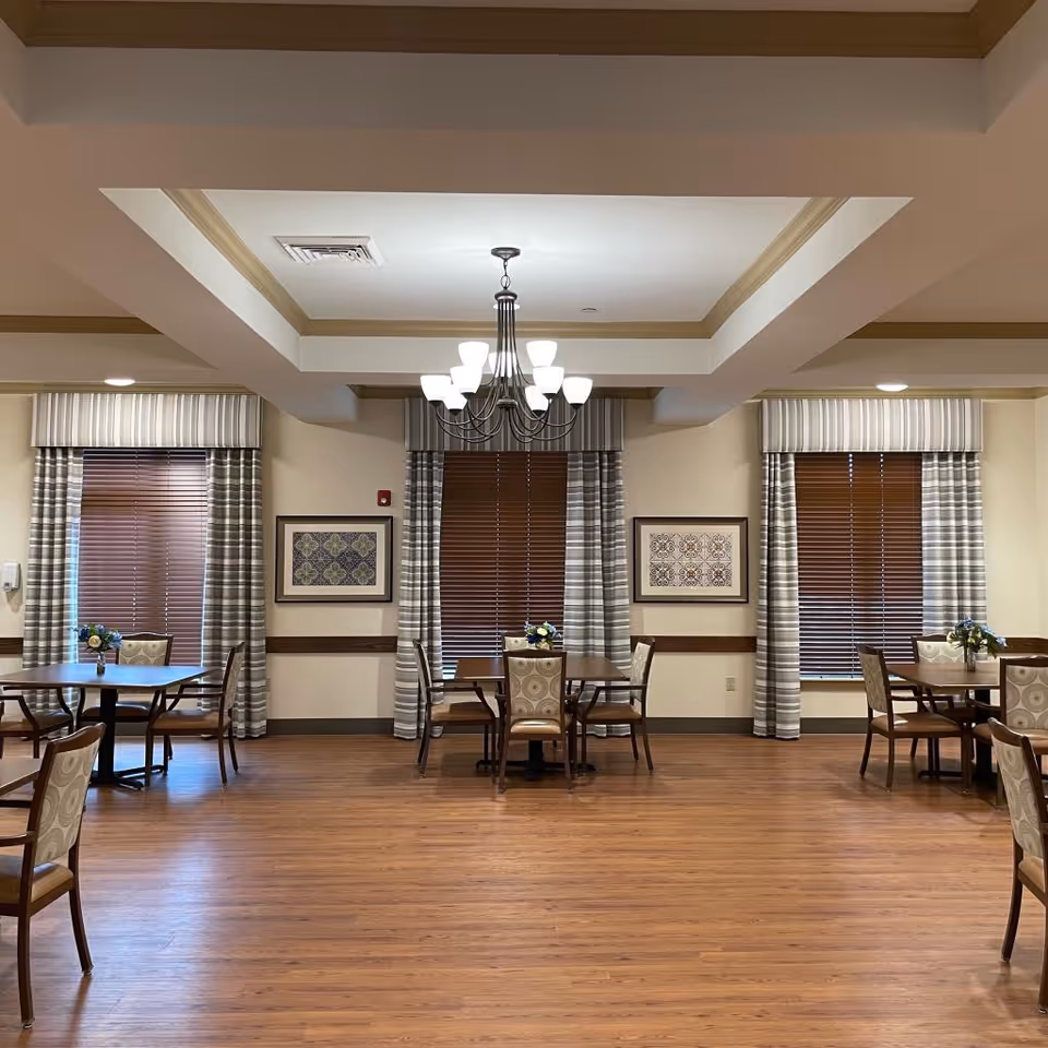A dining room with wooden floors, several tables and chairs arranged neatly, three windows with brown blinds and striped curtains, two framed artworks on the wall, and a chandelier hanging from the ceiling.