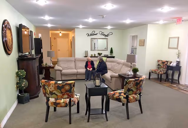 A cozy living room area in a senior living facility with a beige sectional sofa where two elderly women are seated and conversing. The room features two floral-patterned chairs around a small black table, a TV mounted on a wooden stand, decorative plants, lamps, and a wall decoration that says 'Welcome Friends'.