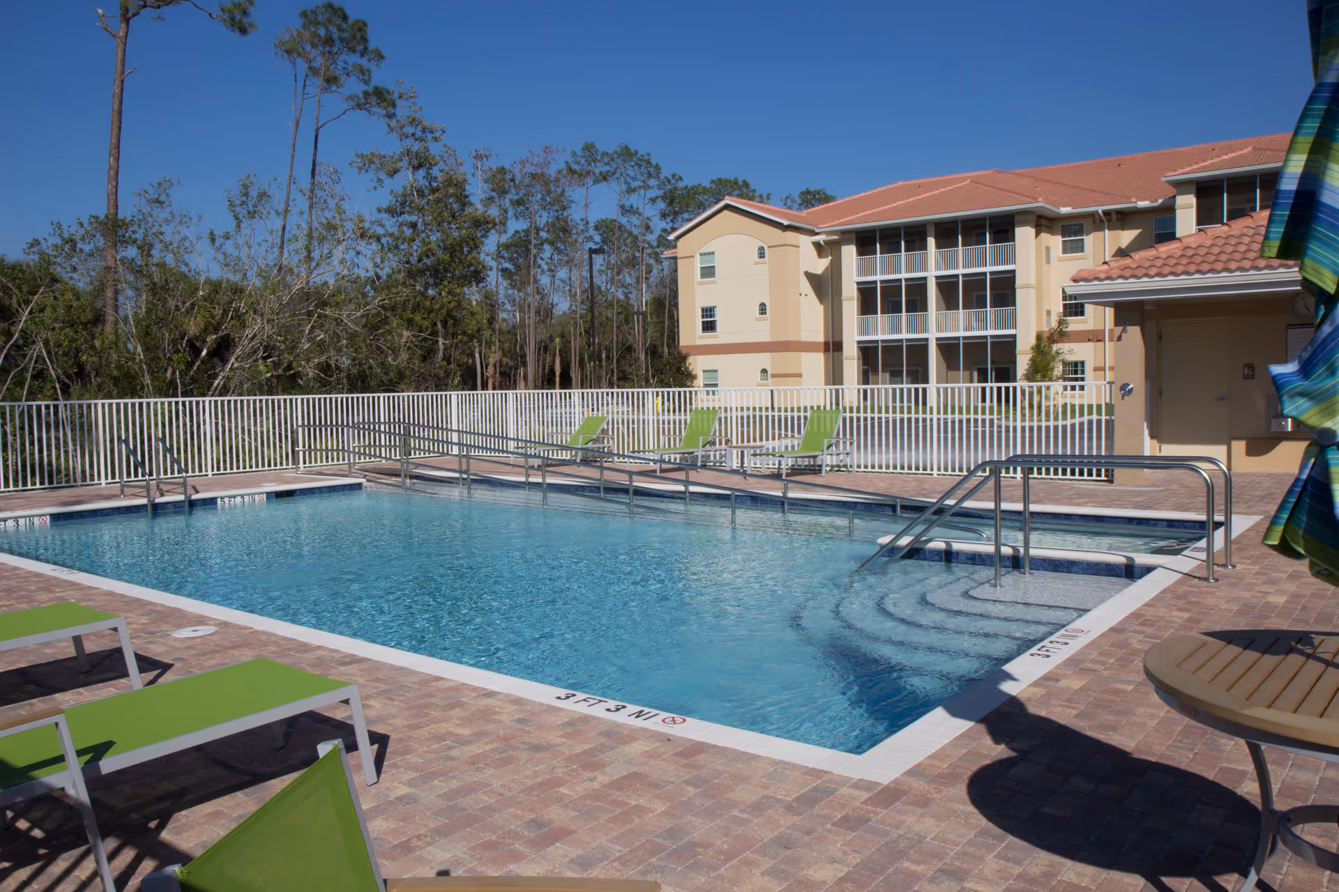 Outdoor swimming pool area with clear blue water, surrounded by a brick-paved deck with green lounge chairs and a round table with a colorful umbrella. In the background, there is a three-story beige building with balconies and a white fence enclosing the pool area. Tall trees and a clear blue sky are visible beyond the fence.