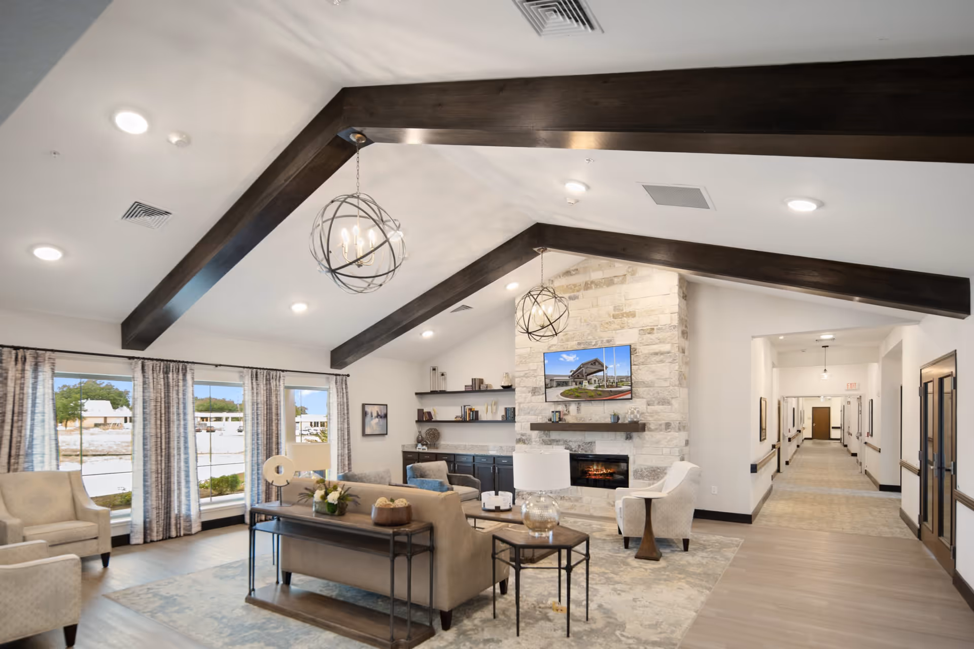 A spacious and well-lit senior living facility lounge area with beige sofas and armchairs arranged around a stone fireplace. Above the fireplace is a mounted flat-screen TV displaying an exterior view of the facility. The room features dark wooden ceiling beams, modern spherical pendant lights, large windows with patterned curtains, and a long hallway leading to other rooms.