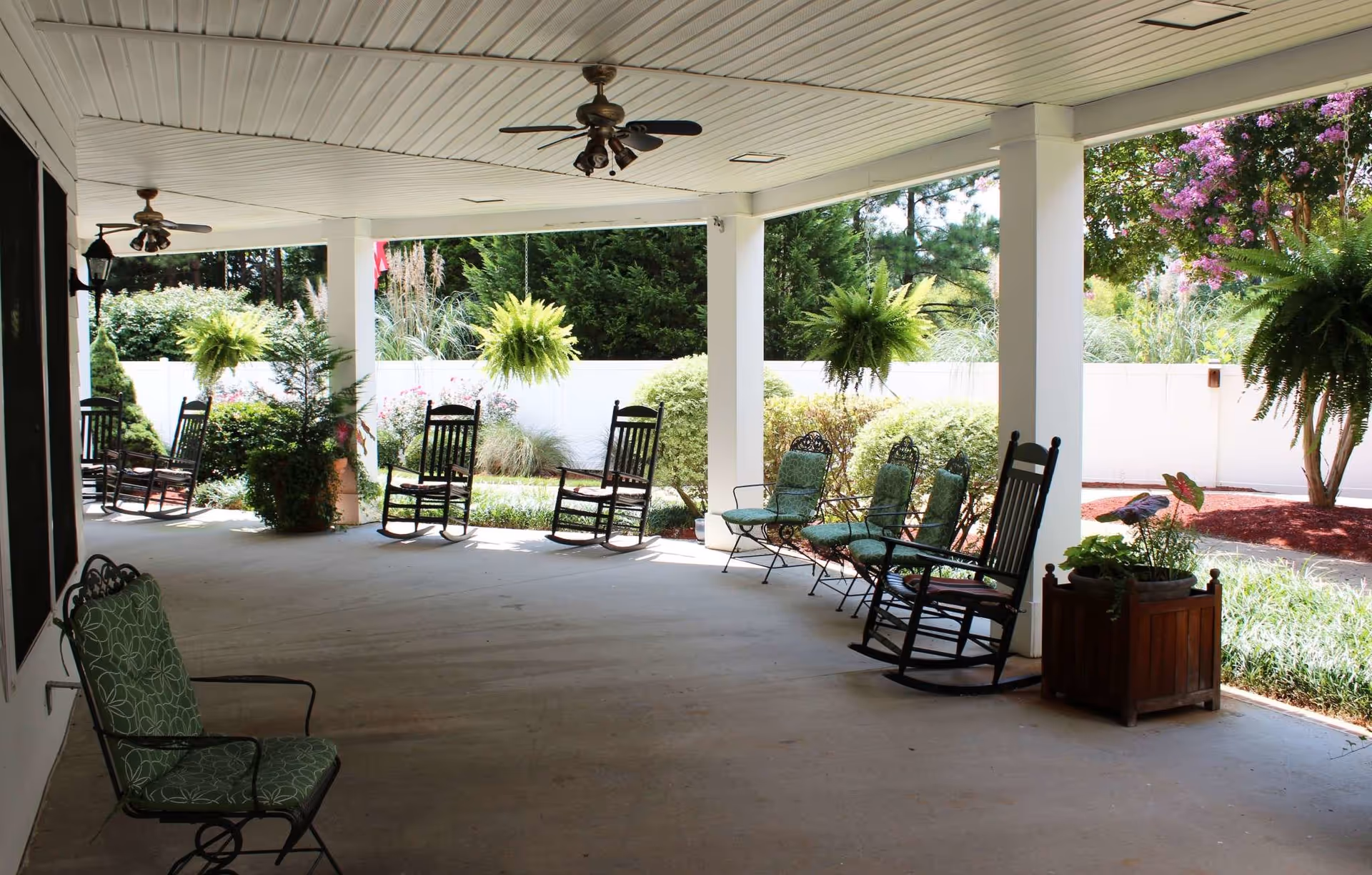 Covered outdoor patio area with several rocking chairs and cushioned metal chairs arranged along the perimeter. Ceiling fans are mounted on the white paneled ceiling. Hanging green ferns and various plants surround the patio, with a white fence and trees in the background.