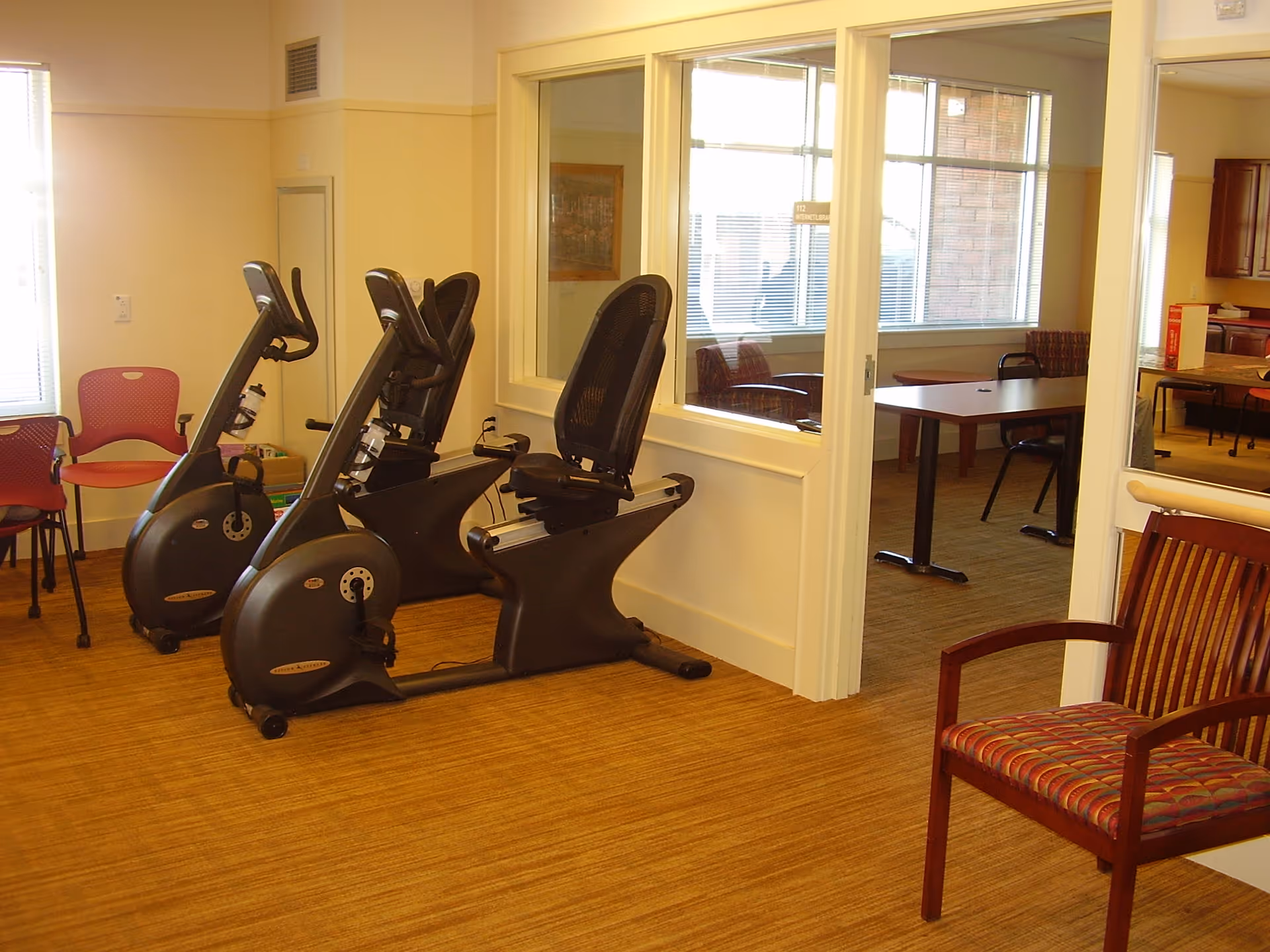 A small exercise room with two stationary recumbent bikes, a red chair, and a wooden chair with a patterned cushion. The room has beige walls and carpeted floor. Through a large interior window, another room with tables and chairs is visible.