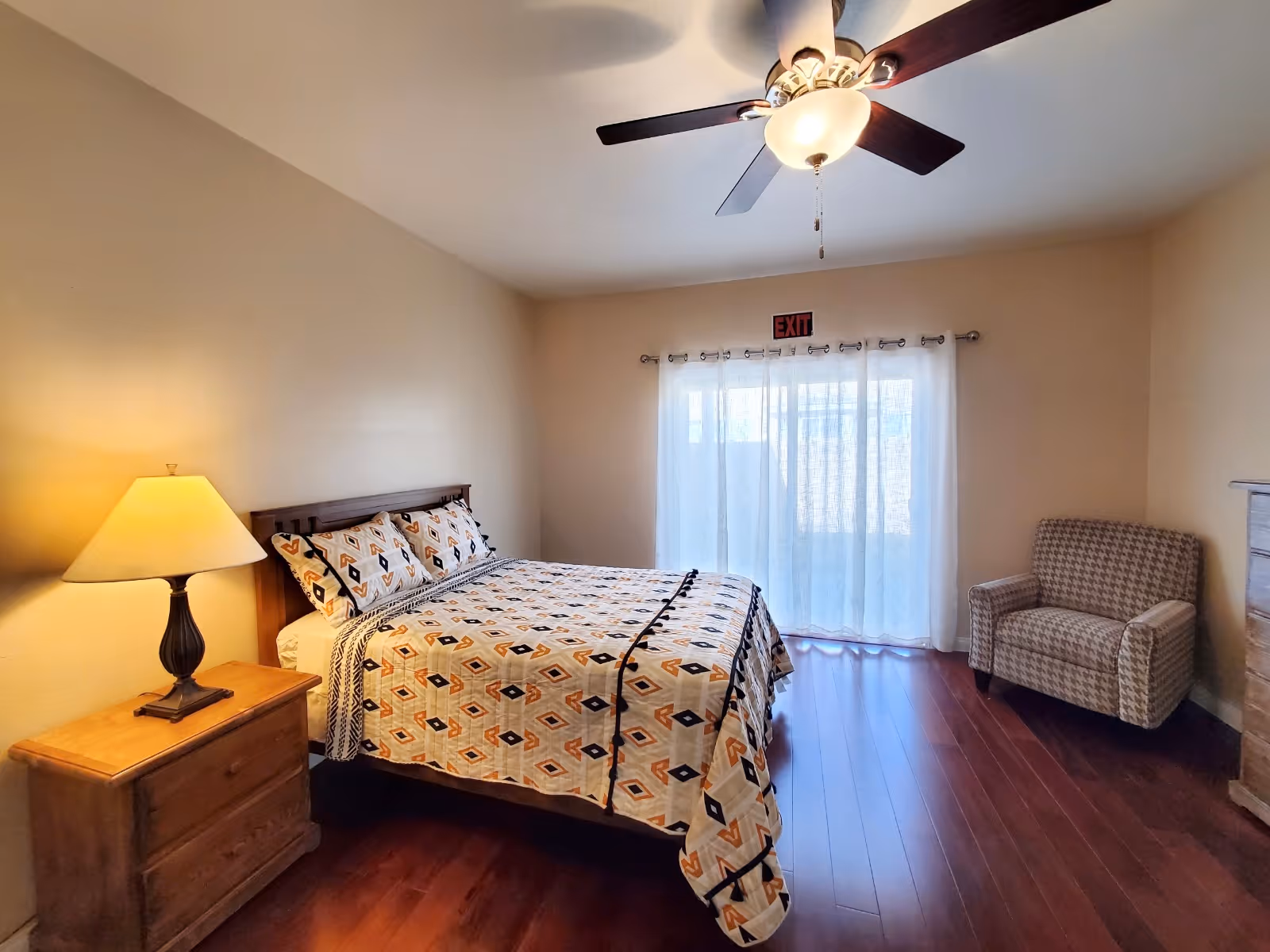 A bedroom with a bed covered in a patterned quilt, a wooden nightstand with a lamp, a cushioned armchair, hardwood floors, a ceiling fan with light, and sheer curtains covering a window with an exit sign above it.