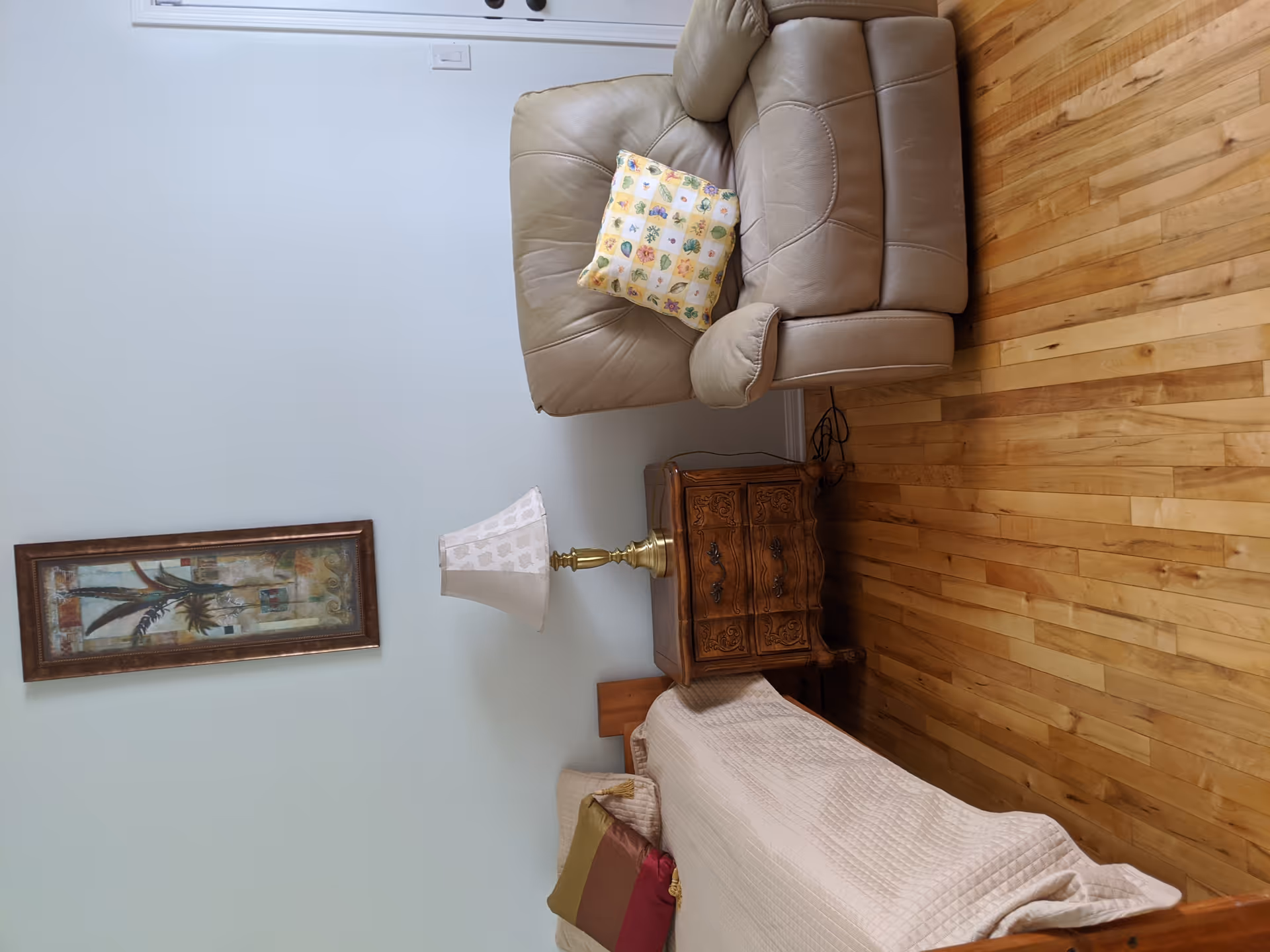 A cozy room with a wooden bed covered in a beige quilt and colorful pillows, a wooden nightstand with ornate carvings holding a brass lamp with a patterned lampshade, a beige leather armchair with a yellow floral cushion, and a framed artwork of plants hanging on a white wall above the nightstand. The floor is made of light-colored wood.