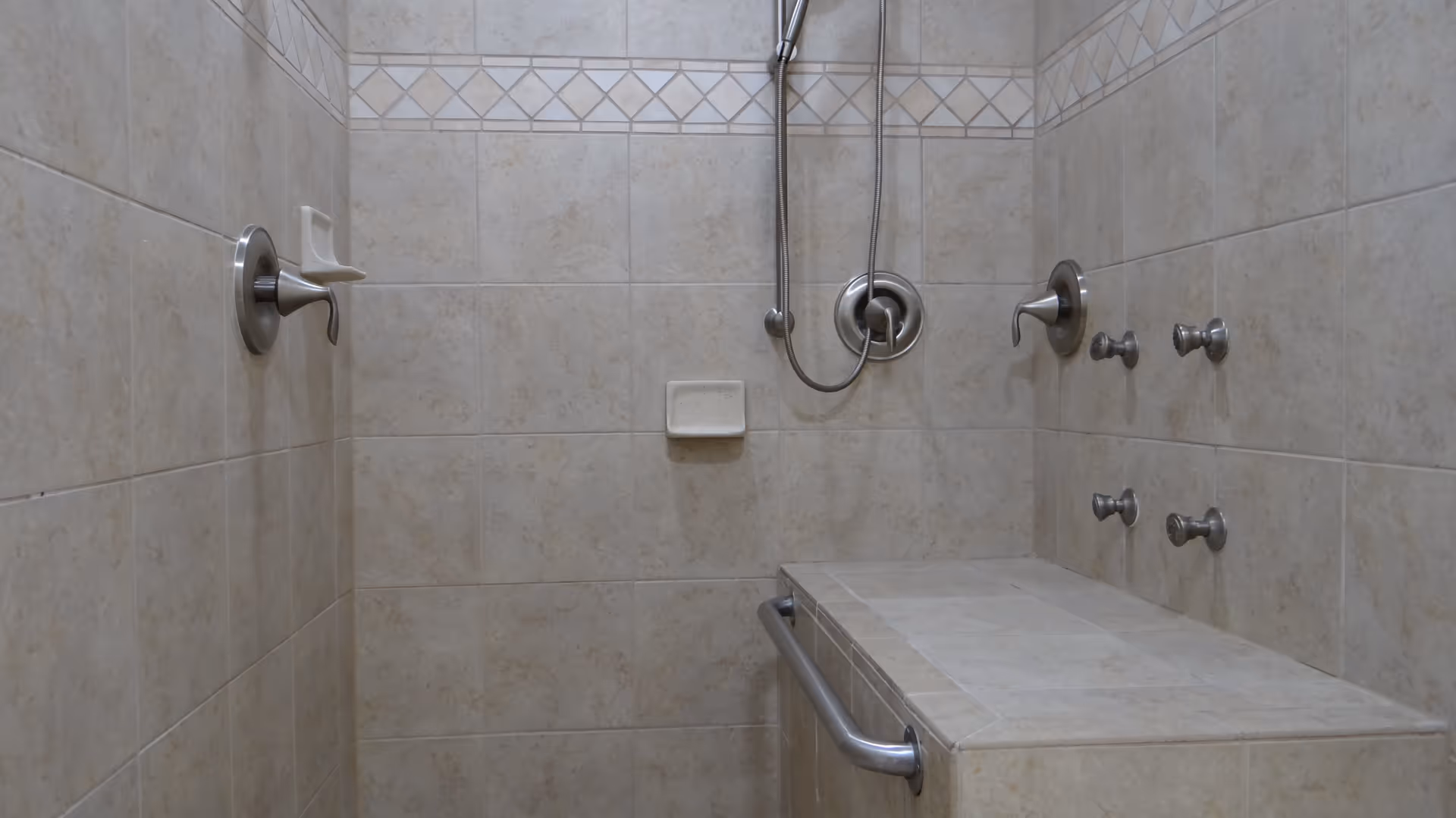 Tile-lined accessible shower stall with a built-in bench, grab bar, handheld showerhead and multiple controls.
