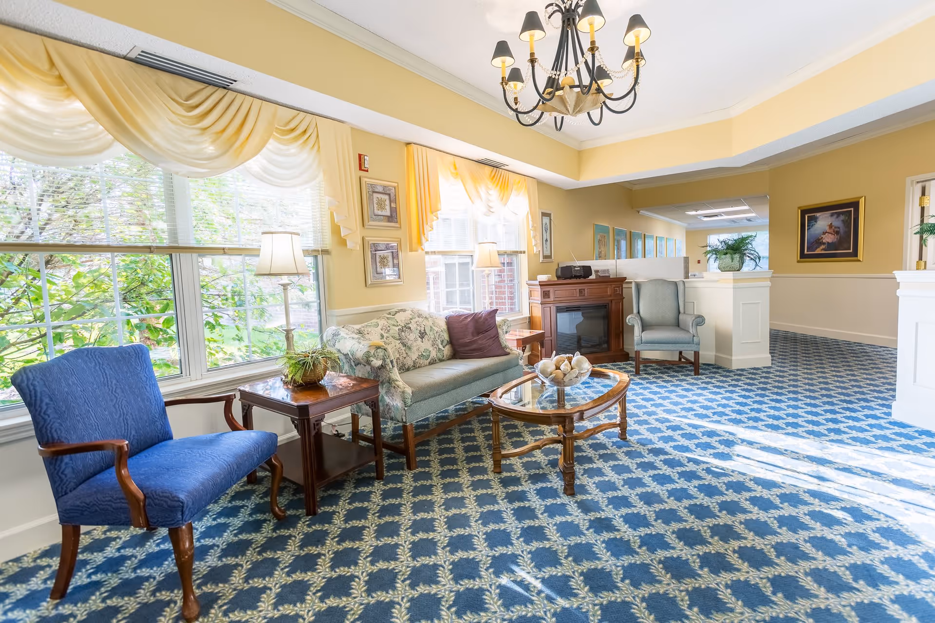 A bright and inviting living room area with large windows covered by sheer yellow curtains, a floral-patterned sofa, two armchairs, a wooden coffee table with decorative items, a side table with a lamp and plant, a chandelier hanging from the ceiling, and framed artwork on the walls. The room has a blue patterned carpet and soft yellow walls.