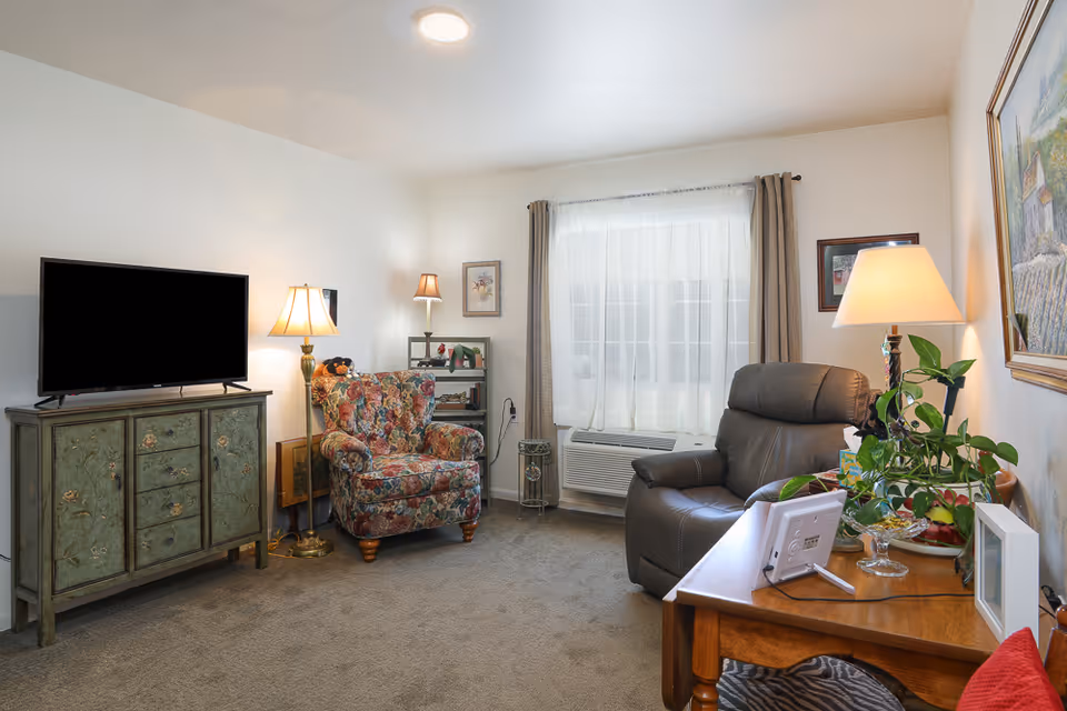 A cozy living room with a floral upholstered armchair, a dark leather recliner, a wooden TV stand with a flat-screen TV, a wooden side table with a lamp and plants, and a window with sheer white curtains and beige drapes.