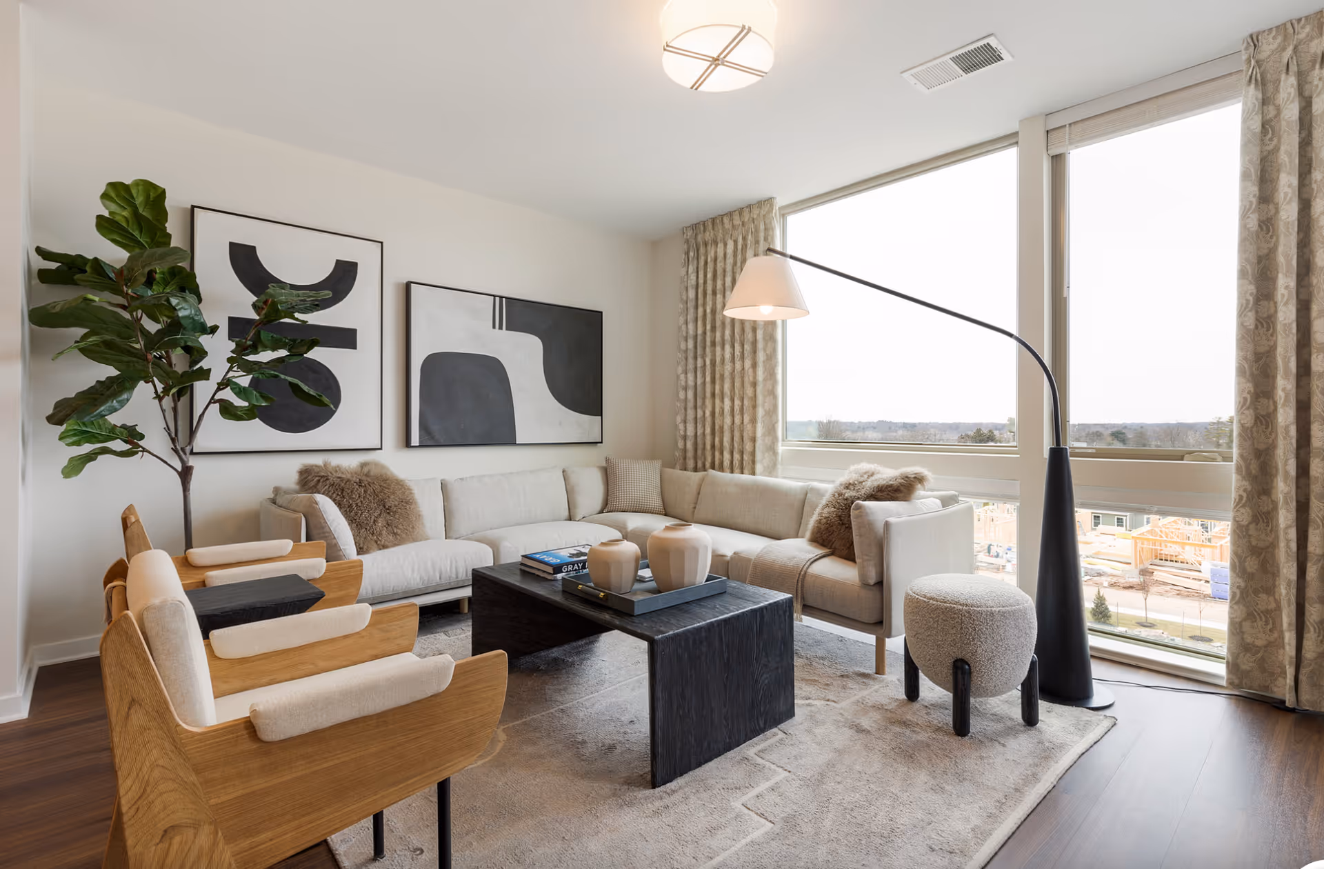 A modern living room with a large beige sectional sofa adorned with cushions and fur throws, two wooden armchairs with light cushions, a black rectangular coffee table with decorative vases and books, a tall black floor lamp with a beige lampshade, a small round upholstered stool, large windows with patterned curtains letting in natural light, and two abstract black and white paintings on the wall next to a tall green plant.