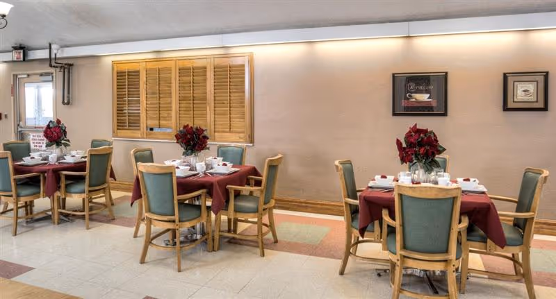 Dining area with three tables covered in burgundy tablecloths, each set with white dishes and decorated with red floral centerpieces. The room has light-colored walls, a window with wooden shutters, framed artwork on the wall, and green cushioned wooden chairs around the tables.